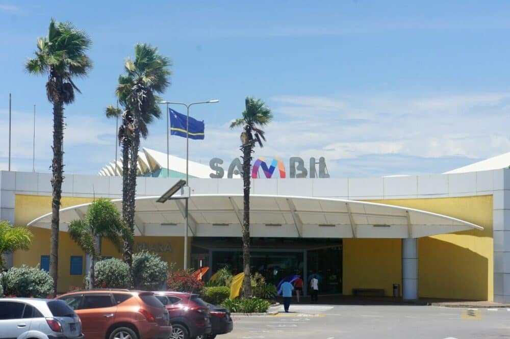 Exterior view of Sambil shopping mall entrance with palm trees and blue sky background.